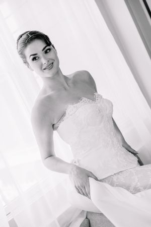 Black and white portrait of a smiling bride in a strapless lace wedding dress posing elegantly near a bright window.