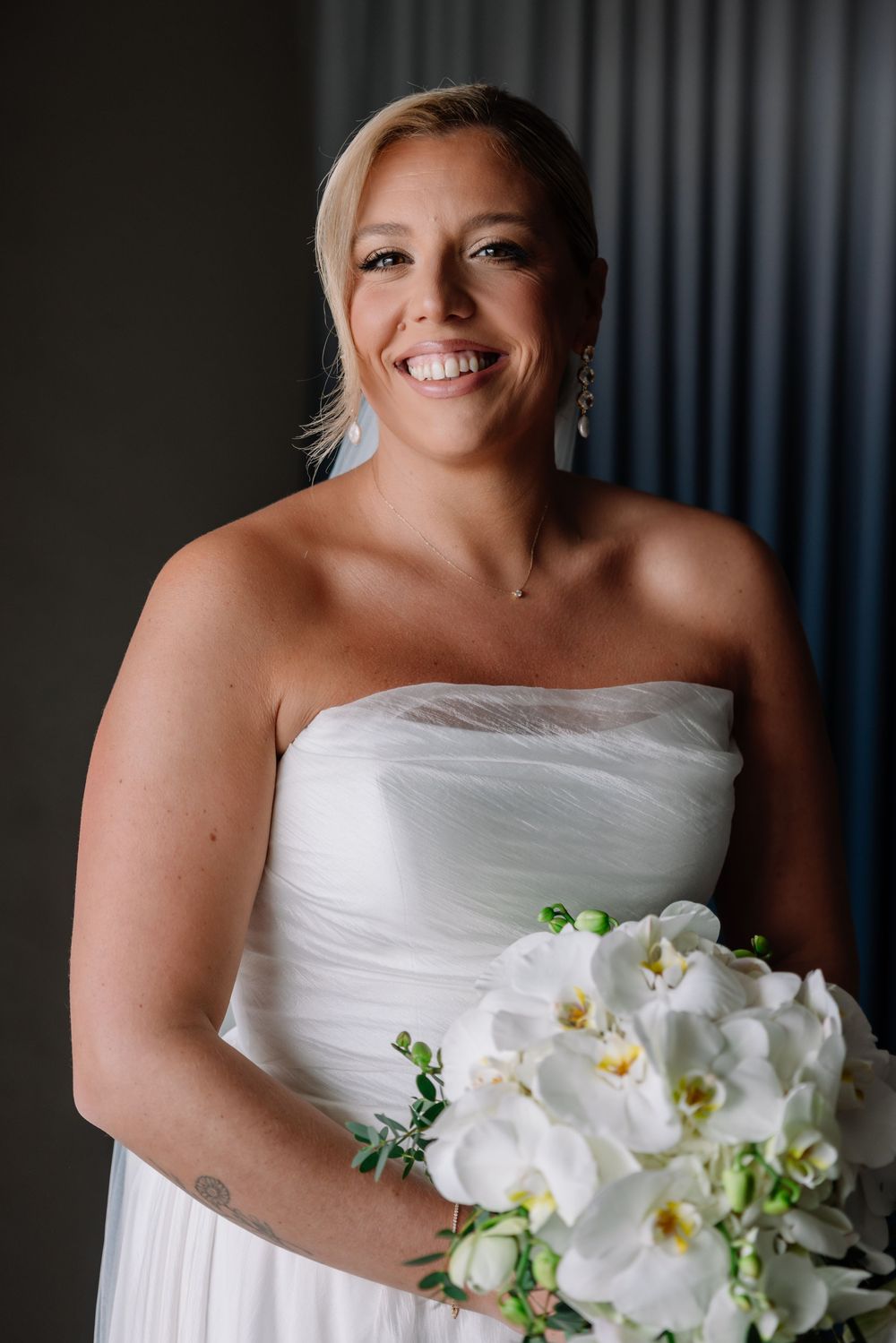 Smiling bride in strapless white gown holding white orchid bouquet, posing indoors on wedding day.