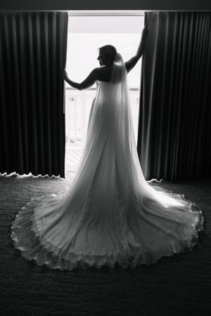 Black and white photo of a bride in a flowing wedding gown with long veil standing at a sunlit window.