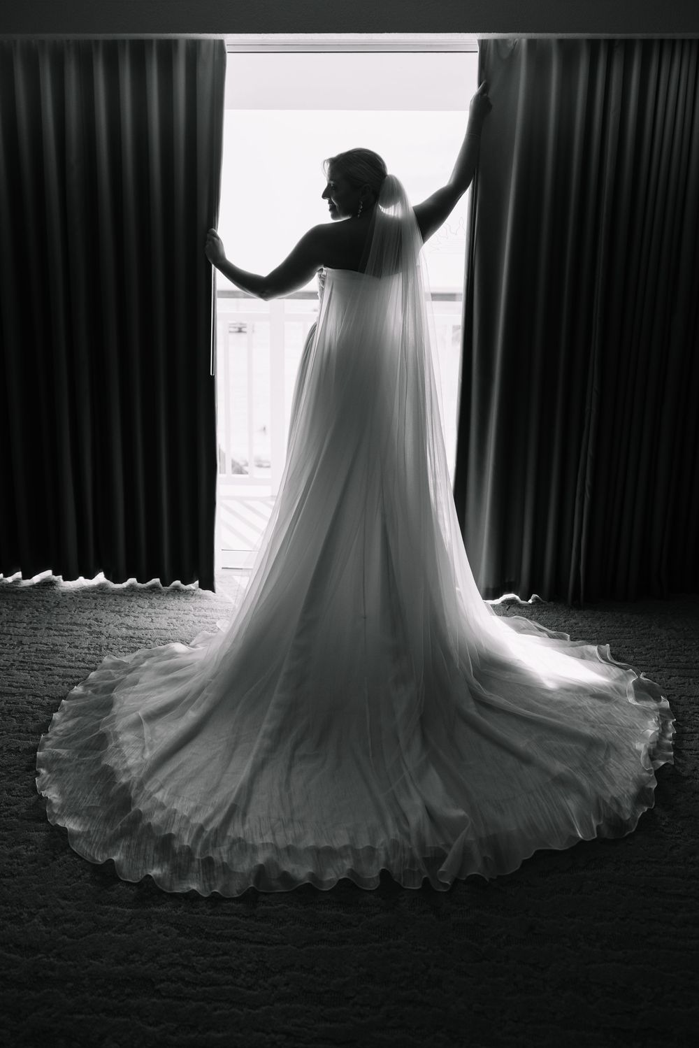 Black and white photo of a bride in a flowing wedding gown with long veil standing at a sunlit window.