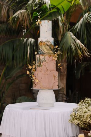 Elegant four-tier wedding cake with gold leaf, blush pink marble, and painted mountain design on a white pedestal stand.
