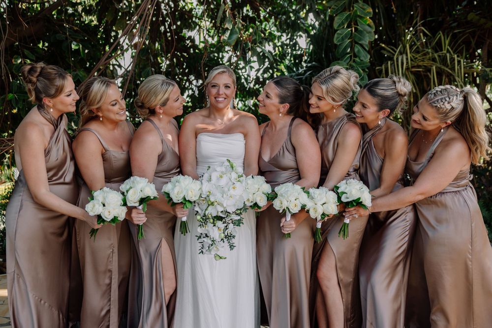 Bride in white gown with bridesmaids in champagne satin dresses holding white floral bouquets outdoors.