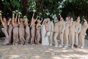 Wedding party in matching champagne attire cheering as bride and groom kiss outdoors amid lush greenery.