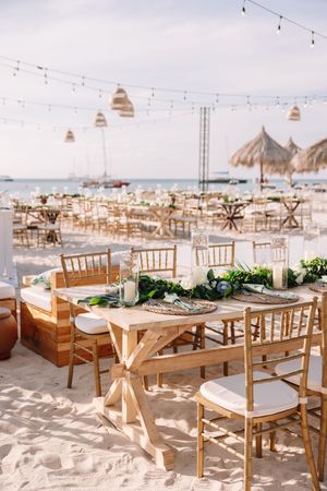Elegant beach wedding reception setup with wooden tables, gold chiavari chairs, greenery centerpieces, string lights, and ocean views.