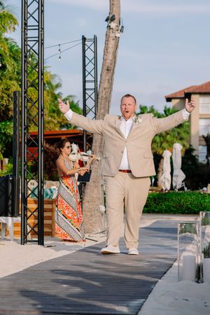 Excited groom in tan suit walks down outdoor aisle with arms raised at tropical beach wedding venue.