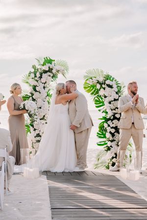 Bride and groom share first kiss at beach wedding altar adorned with white flowers and tropical leaves.