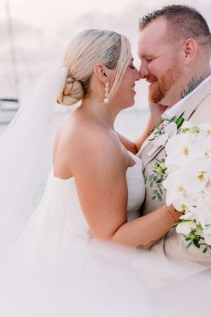 Bride and groom share an intimate moment, touching foreheads and smiling, bride holding white orchid bouquet in wedding attire.