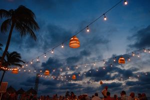 Outdoor evening event with string lights and glowing lanterns under a dramatic blue cloudy sky near tropical palm trees.