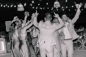 Black and white photo of wedding guests dancing joyfully under string lights at an outdoor evening reception.