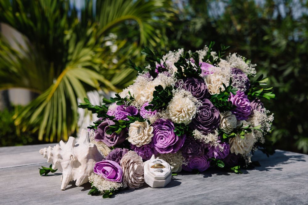 Purple and white wedding bouquet with roses and dahlias beside seashells on a table with tropical greenery.