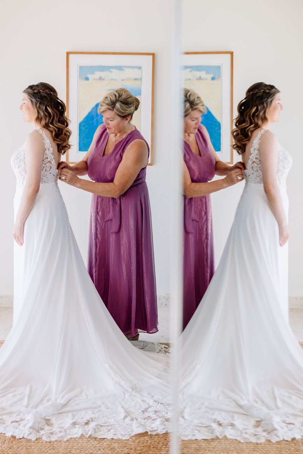 A bride in a white lace gown gets her dress fastened by a woman in purple, reflected in a mirror.