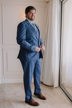 A smiling man in a blue suit with brown dress shoes stands near a window, buttoning his jacket in an elegant room.