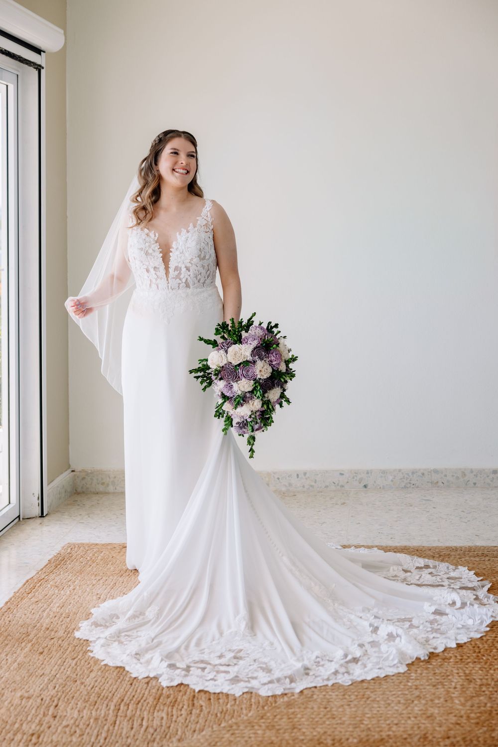 Bride in white lace V-neck gown with train holds purple and white bouquet, smiling near window.