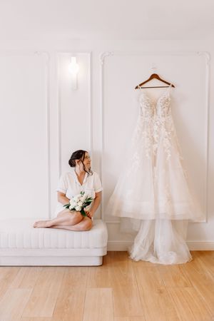 Bride in white robe sitting on bench holding bouquet beside hanging lace wedding dress in bright white room.