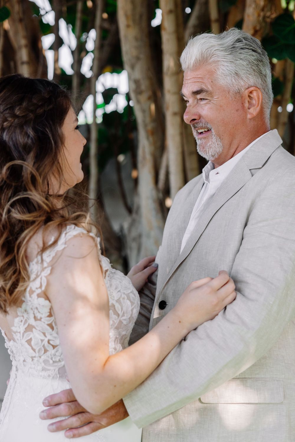 Bride in lace wedding dress shares tender first look moment with father in gray suit outdoors.