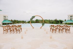 Outdoor wedding ceremony setup with wooden chairs, circular gold arch, candles, and ocean view at a tropical resort.