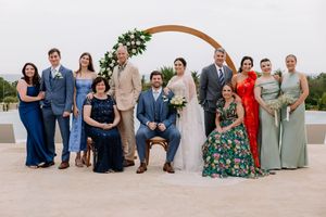 Wedding party poses in front of floral arch, bride and groom centered with family in formal attire outdoors.