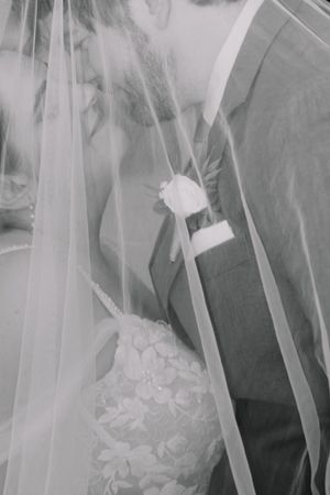 Black and white wedding photo of bride and groom kissing under veil, groom wearing rose boutonniere.