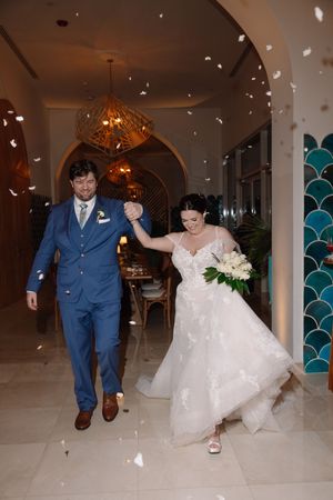 Bride and groom walk through confetti shower, she in white gown with bouquet, he in blue suit, at elegant venue.