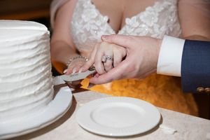 Bride and groom cutting white wedding cake together, bride wearing diamond engagement ring, elegant reception setting.
