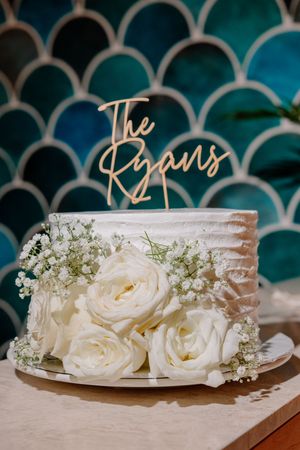 Elegant single-tier wedding cake with white roses, baby's breath, and gold "The Ryans" cake topper on teal tile background.