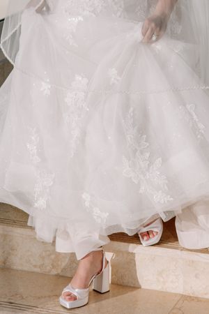 Bride lifting her white lace wedding gown to reveal elegant white platform heels on marble steps.