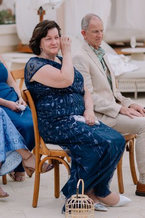Emotional woman in blue dress wiping tears at wedding ceremony seated beside man in tan suit.