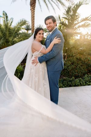 Bride in lace gown with flowing veil embraces groom in blue suit outdoors with palm trees and golden sunset.