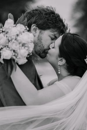 Black and white photo of a bride and groom sharing a romantic kiss, with a white floral bouquet and veil flowing.