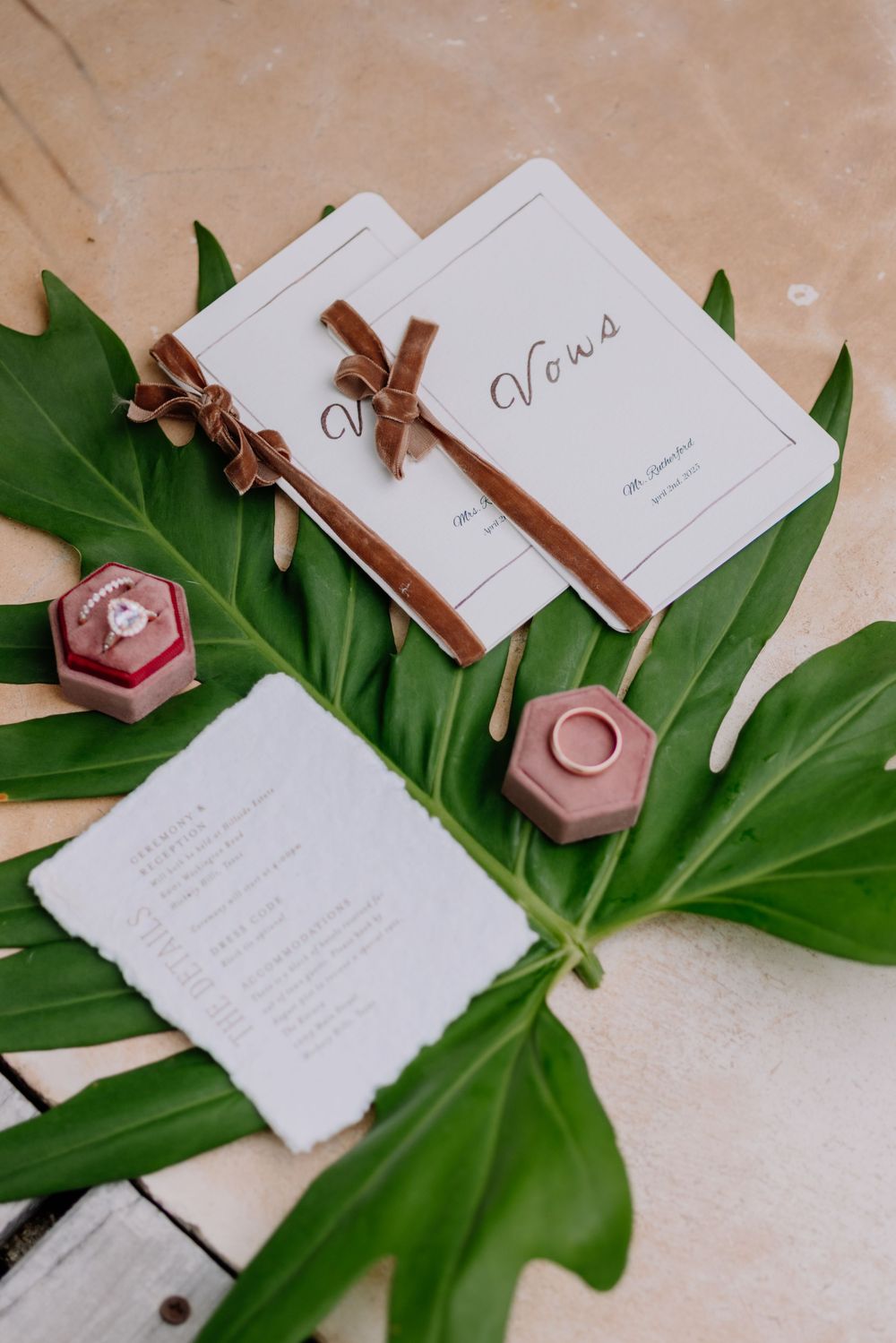 Elegant wedding vows booklets with brown velvet ribbons, ring boxes, and tropical leaf flat lay arrangement.