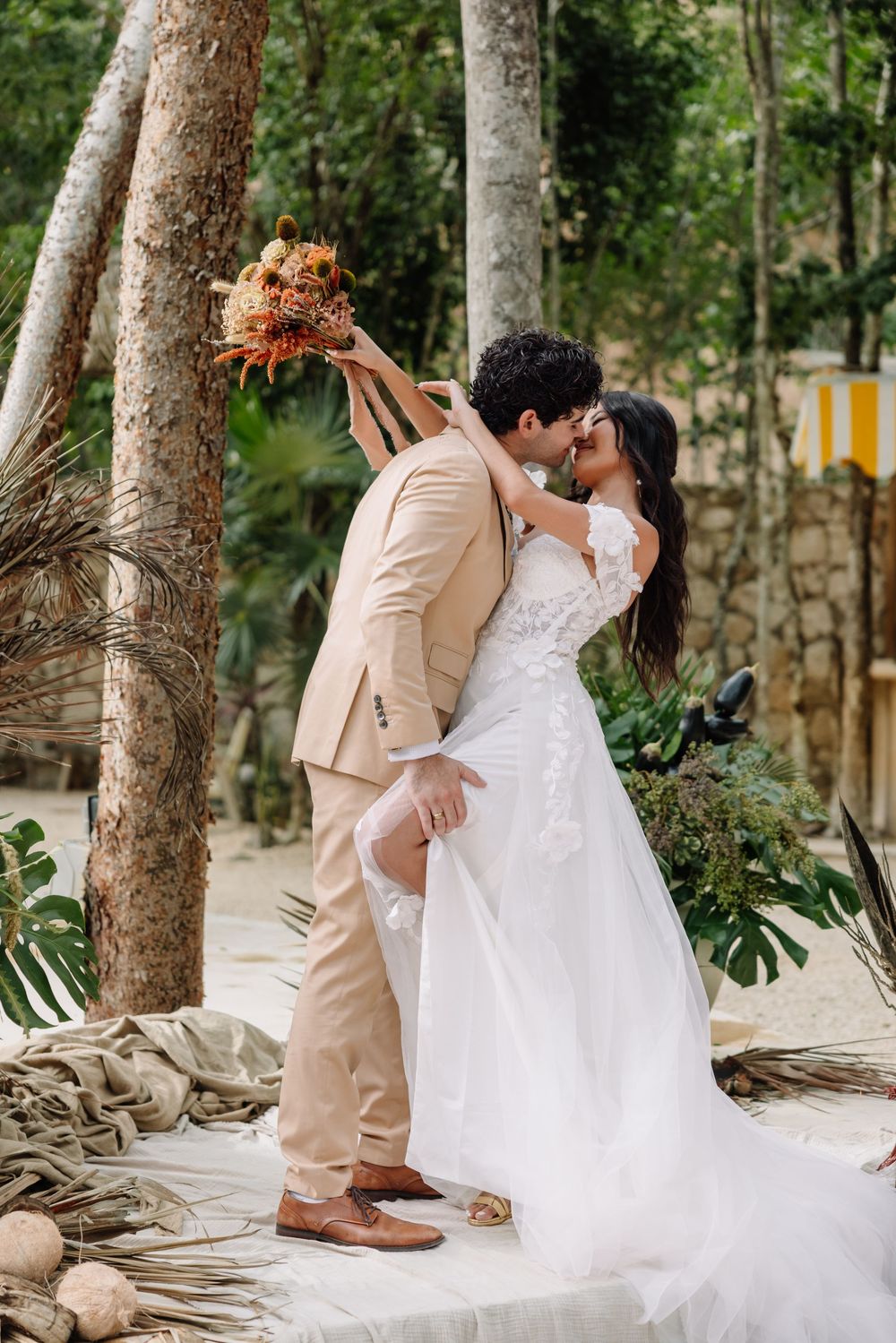 Bride and groom share a romantic kiss outdoors in a tropical forest setting, with the bride in a white gown holding a bouquet.
