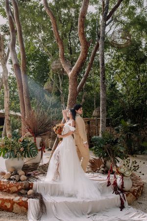 Bride and groom stand on outdoor wedding altar surrounded by lush trees, tropical plants, and boho decor.