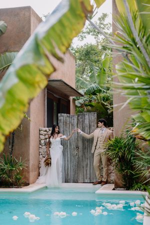 Bride and groom hold hands by a tropical pool, framed by lush palms and rustic wooden gates at an outdoor wedding venue.