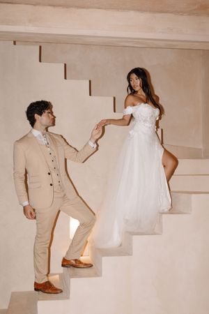 Bride in white off-shoulder gown holds groom's hand on elegant stairs, groom in beige suit.