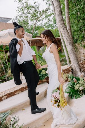 Joyful interracial couple in wedding attire holding hands on stone steps surrounded by tropical greenery.
