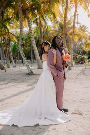 Interracial couple in wedding attire on sandy beach with palm trees, bride in white gown, groom in pink suit holding bouquet.