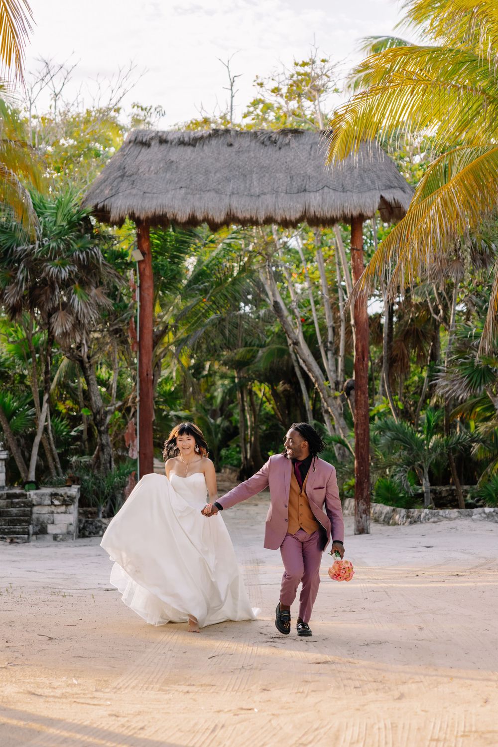 Bride and groom walk hand-in-hand on sandy beach with tropical palms and thatched gazebo at destination wedding.