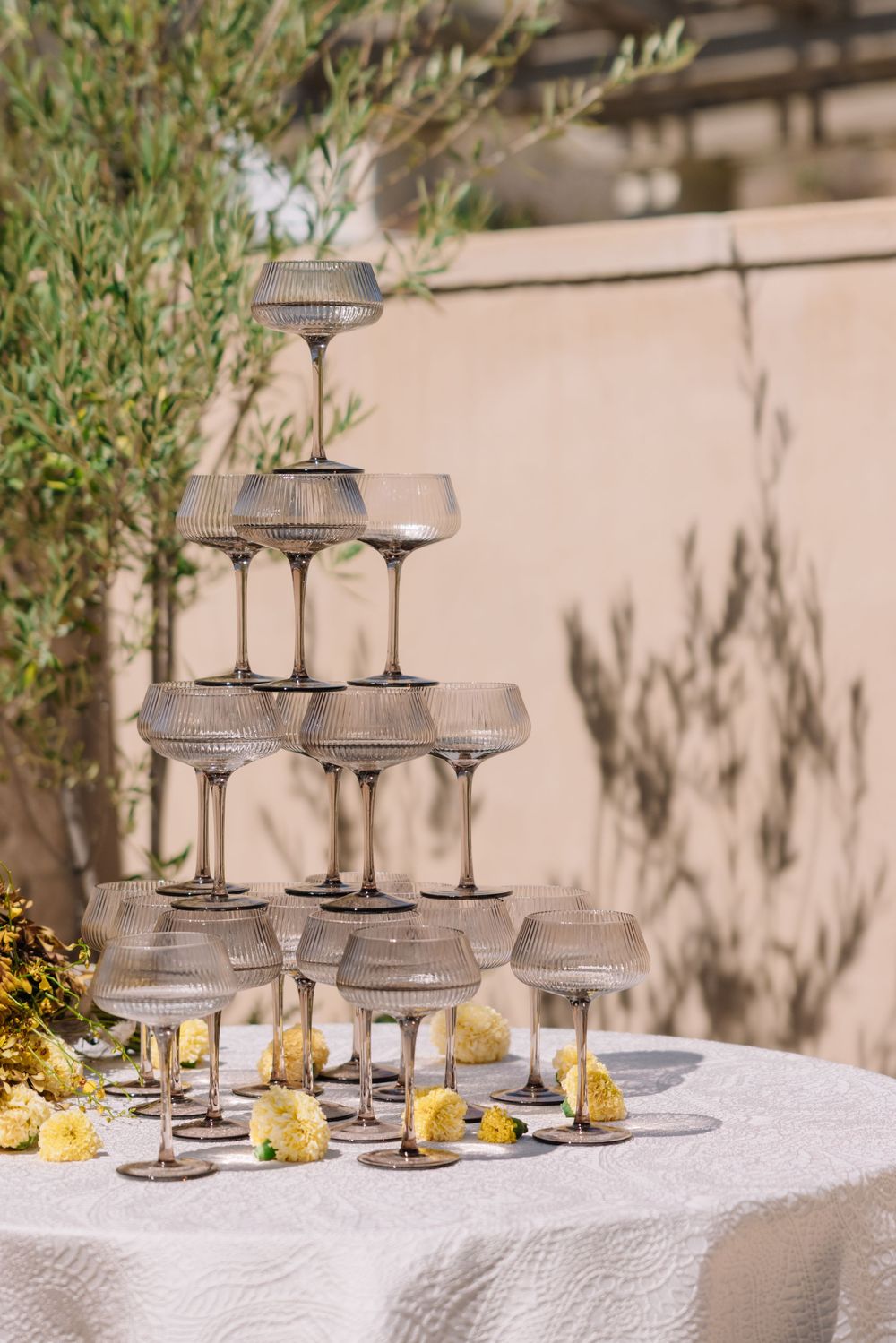 Elegant champagne tower with ribbed coupe glasses on a white lace table outdoors, decorated with yellow flowers.