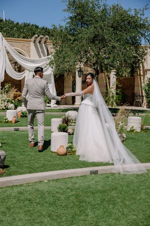 Bride in white gown and groom in gray suit hold hands in lush garden wedding venue with draped canopy backdrop.