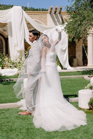 Bride and groom stand back-to-back outdoors, bride lifting her flowing white veil at an elegant garden wedding venue.