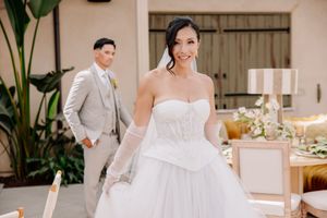 Bride in strapless white ball gown with gloves smiles outdoors as groom in gray suit stands in background.