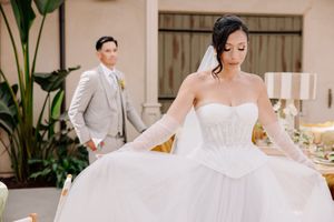 Bride in white strapless ballgown with gloves holds dress while groom in gray suit stands in background.