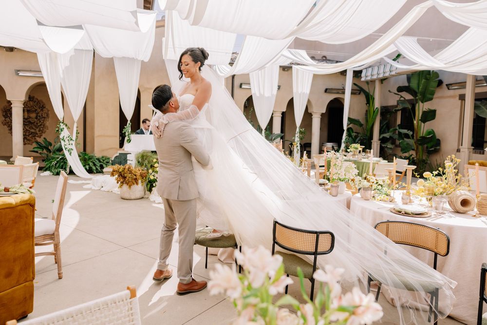 Groom lifts bride in white gown and flowing veil at elegant outdoor wedding reception with draped fabric ceiling.