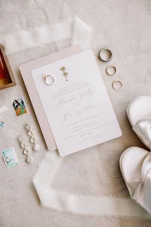 Wedding flat lay featuring invitation suite, gold rings, pearl earrings, bridal shoes, and ribbon on neutral linen.