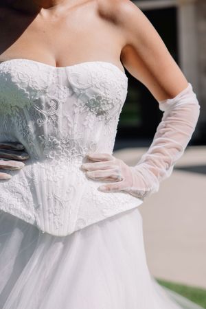 Bride in strapless white lace corset wedding gown with sheer gloves, hands on hips, showcasing intricate floral embroidery.