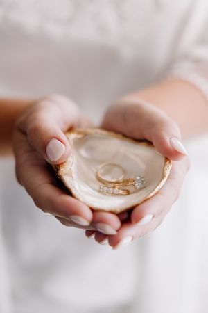 Hands holding an oyster shell ring dish with gold wedding bands and a diamond engagement ring.