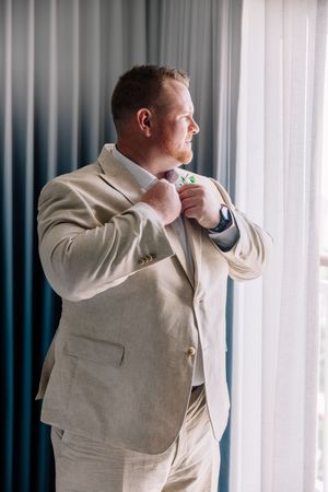 Groom in beige linen suit adjusting his tie near a window with blue curtains on his wedding day.