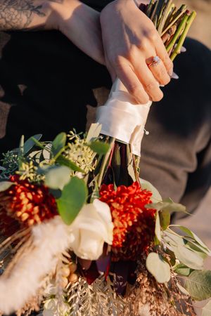 Close-up of bride holding a wedding bouquet with red and white flowers, greenery, and white ribbon, wearing an engagement ring.