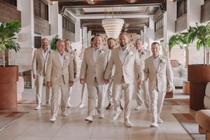 Groom and groomsmen in matching beige suits walking confidently through an elegant resort lobby.