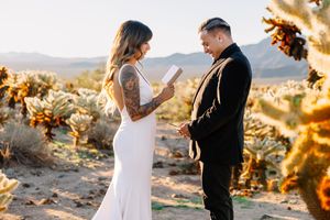 Couple exchanging vows in desert wedding ceremony among Joshua trees, bride in white gown with tattoos, groom in black suit.